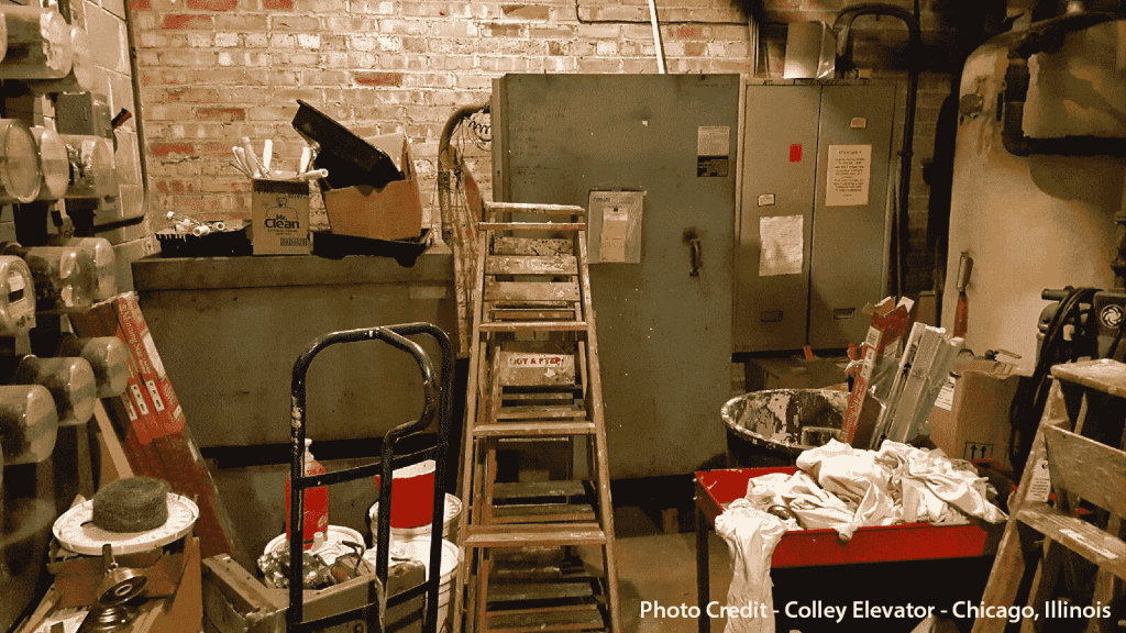 Cluttered elevator machine room with tools, ladders, boxes, and electrical panels, showing poor maintenance conditions