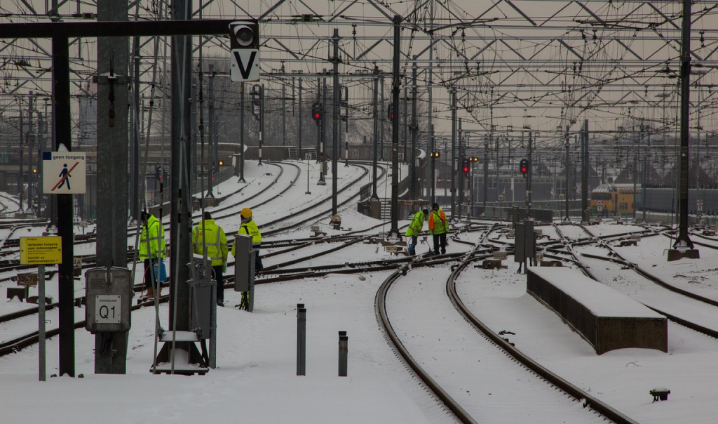 Railway workers in high-visibility jackets inspect and clear snow from multiple train tracks at a snowy rail yard, with overhead power lines and red signal lights visible in the background.