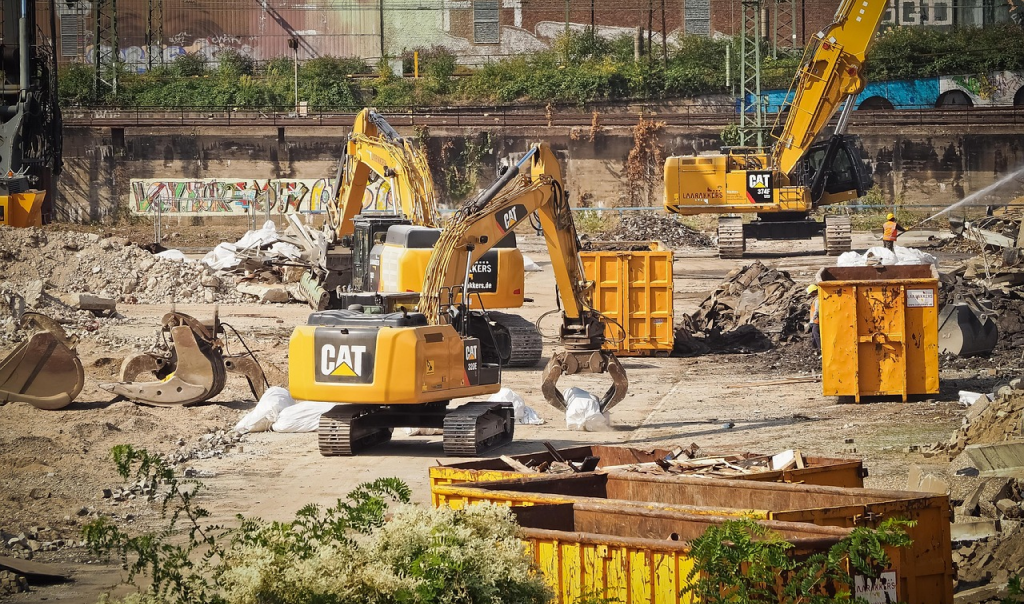 Several yellow CAT excavators operating at a demolition site, surrounded by rubble and debris, with large metal dumpsters and a worker spraying water in the background near a graffiti-covered wall.