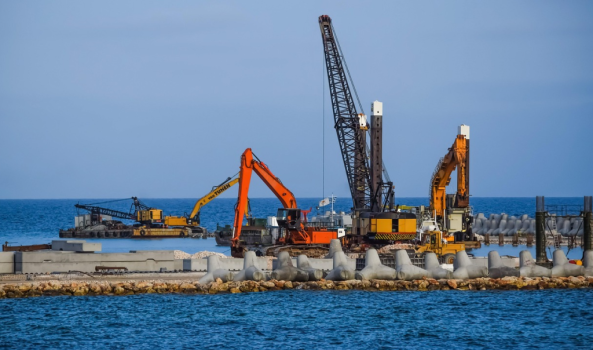 An industrial scene at a coastal construction site under a clear blue sky. Several pieces of heavy machinery, including large orange and yellow excavators and a tall lattice boom crane, are positioned on a rocky breakwater and a barge in the water. In the foreground, a line of grey concrete tetrapods (four-legged breakwater structures) sits along the edge of the blue sea, intended for coastal protection. The water is calm, and additional construction materials and concrete blocks are visible on the site.