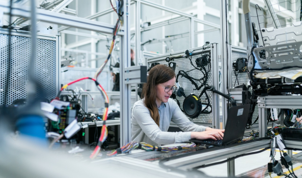 A woman with long brown hair and glasses sits at a workbench in a brightly lit industrial or laboratory setting. She is focused on a laptop, with her hands on the keyboard. The environment is filled with complex machinery, aluminum framing, and intricate bundles of colorful wiring and electronic components, suggesting a high-tech manufacturing or engineering workspace.