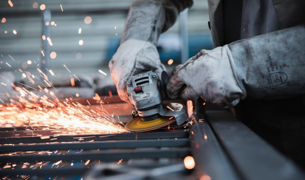 A person wearing protective gloves uses an angle grinder to cut or smooth metal, sending bright sparks flying across a workshop surface.