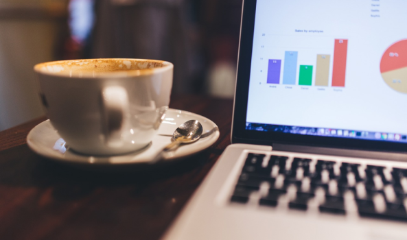 A cup of coffee on a saucer with a small spoon sits on a table next to an open laptop displaying colorful charts and graphs on the screen.