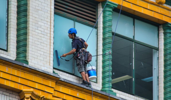 A worker wearing a safety helmet and harness is suspended by ropes while cleaning a large window on the exterior of a colorful building, using a squeegee and carrying a bucket attached to his gear.