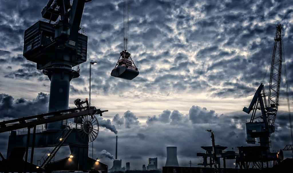 Industrial port scene at dusk with large cranes lifting cargo, smokestacks emitting steam in the background, and dramatic cloud-filled sky over a working harbor.